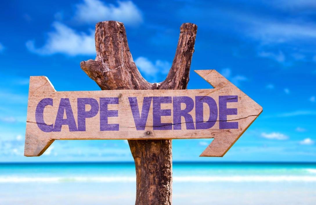 A wooden sign displaying "Cape Verde" stands on a sandy beach, surrounded by gentle waves and clear blue skies.